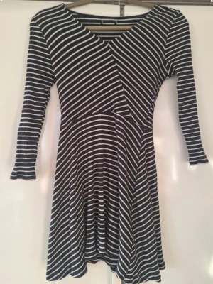 Randig omlottklänning med trekvartsärm - Mörkblå och vit randig kortklänning med omlott framtill och v-ringning. Klänningen har trekvartsärm och är tillverkad i mjukt, stretchigt material som ger en skön passform. Perfekt för dig som gillar en avslappnad men ändå snygg stil.