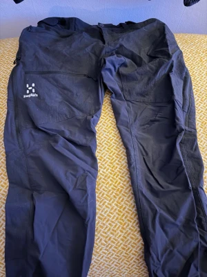 Svarta cargobyxor från Haglöfs - Säljer ett par svarta cargobyxor från Haglöfs med flera praktiska fickor och dragkedja på sidan. Byxorna har en sportig och funktionell design, perfekta för dig som gillar outdoor och streetwear. Materialet känns slitstarkt och byxorna har en modern passform. Står Men 50 men jag upplever dom som S-M