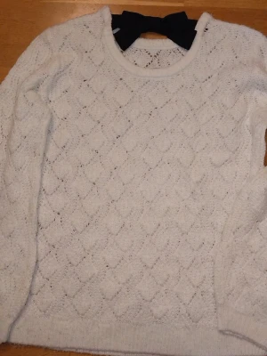 Vit stickad tröja med rosett - Supersöt vit stickad tröja från Lindex med ett romantiskt hålmönster och en stor svart rosett i nacken. Tröjan har rund halsringning och långa ärmar. Perfekt för dig som gillar en gullig och trendig stil. För barn 10-12 men passar även 13. Priset kan diskuteras❤️ 