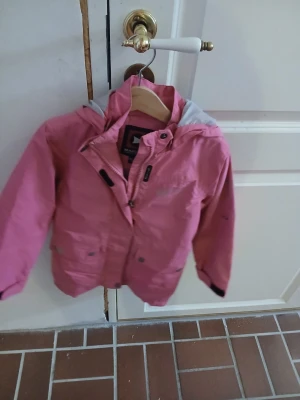 Rosa vindjacka med huva från marine - Snygg rosa vindjacka från marine med grått foder i huvan. Jackan har dragkedja, tryckknappar och två fickor framtill. Perfekt för blåsiga dagar och har en sportig look med resår i midjan och reflexdetalj bak.