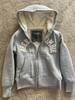 Grå hoodie från Abercrombie & Fitch - Mysig grå hoodie från Abercrombie & Fitch med dragkedja och vit teddyfoder på insidan. Stor logga och siffrorna 1892 broderat framtill. Storlek Small! Men sitter som Xs då den är mindre än S!Pris kan diskuteras!