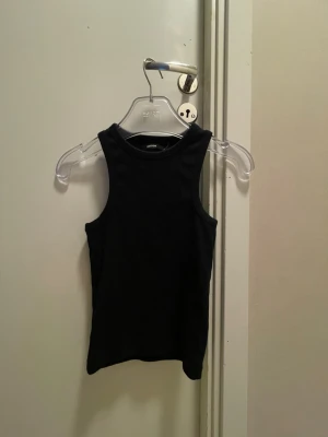 Svart ribbad linnetopp BikBok - Snygg svart ribbad linnetopp från BikBok. Modellen är ärmlös med hög halsringning och tight passform, perfekt att styla till jeans eller kjol. Enkel och stilren design som funkar till många olika looks.