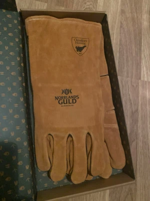 GRILLHANDSKAR!!! - Säljer ett par långa, bruna mockahandskar från Orrefors Hunting med tryck från Norrlands Guld. Handskarna har klassisk femfingermodell och är tillverkade i mjukt mockaläder, perfekta för dig som gillar outdoor och stilrena accessoarer.