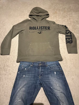 Olivgrön hoodie från Hollister - Snygg olivgrön hoodie från Hollister med stor logga på bröstet och ärmen. Klassisk känguruficka framtill och justerbar huva med dragsko. Tillverkad i mjuk bomullsblandning som är perfekt för chill dagar. Oversized passform och ribbade muddar vid ärmslut och nederkant.
