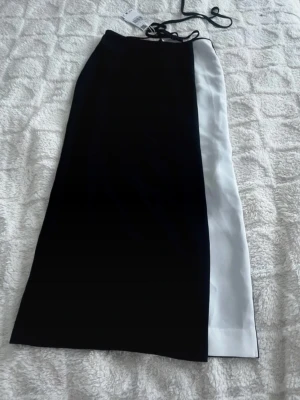 Svart och vit midilång kjol - Snygg midilång kjol med svart bas och en vit panel på ena sidan. Kjolen har rak passform och knytband i midjan för justerbar passform. Perfekt för dig som gillar stilrena kontraster och modern design.