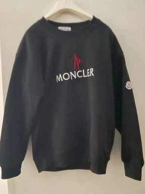 Svart sweatshirt från Moncler - Säljer en svart sweatshirt från Moncler med vit och röd logga broderad på bröstet och ett Moncler-märke på ärmen. Tröjan har rund halsringning och ribbade muddar vid ärmslut och nederkant. Perfekt för en clean och stilren look.