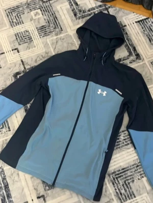 Blå vindjacka från Under Armour - Snygg vindjacka från Under Armour i två nyanser av blått med huva och dragkedja framtill. Jackan har långärmade ärmar och logga på bröstet. Perfekt för dig som gillar sportig stil och vill ha något lätt och funktionellt. Ett litet hål vid armen som inte syns vid användning!