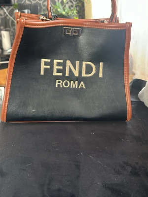 Fendi Roma svart handväska - Cool svart handväska från Fendi med bruna läderdetaljer och guldtext 'FENDI ROMA' på framsidan. Väskan har metallspännen och både handtag och axelrem, perfekt för att bära på olika sätt. Materialet är läder och designen är boxig och stilren. Inte äkta 
