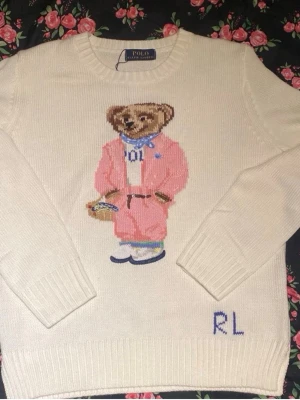 Vit stickad tröja Polo Bear RL - Vit stickad tröja från Polo Ralph Lauren med den ikoniska Polo Bear i rosa outfit på bröstet och blå RL-broderi vid nederkanten. Tröjan har rund hals, långa ärmar och ribbade muddar. Perfekt för dig som gillar klassisk preppy stil.
