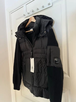 Svart pufferjacka från C.P. Company - Säljer en svart pufferjacka från C.P. Company med stickade ärmar och huva. Jackan har dragkedja framtill, två sidofickor med dragkedja och en ikonisk linsdetalj på ena ärmen. Perfekt för kalla dagar och riktigt snygg streetstil. Den är helt oanvänd. 