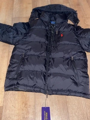 Svart pufferjacka från Polo Ralph Lauren - Säljer en svart pufferjacka från Polo Ralph Lauren med huva och dragkedja framtill. Jackan har två snedställda fickor med dragkedja och den klassiska röda loggan broderad på bröstet. Perfekt för kalla dagar och riktigt snygg till streetwear-stilen.