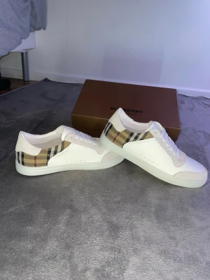 Burberry vita sneakers med rutigt mönster - Snygga vita sneakers från Burberry med det klassiska beige, svarta och röda rutmönstret på sidan. Skorna har vita snören, diskreta hål för ventilation och detaljer i mocka. Perfekta för dig som vill ha en clean och lyxig look.