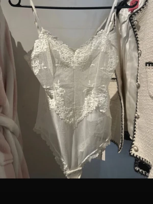 Vit spetsbody med tunna axelband - Säljer en superfin vit body med spetsdetaljer och tunna axelband. Bodyn har v-ringning, meshpartier och dekorativa spetsmönster framtill och längs kanterna. Perfekt för dig som gillar romantisk och feminin stil. Materialet är genomskinligt mesh och spets.