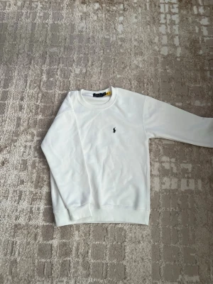 Vit sweatshirt från Polo Ralph Lauren - Klassisk vit sweatshirt från Polo Ralph Lauren med rund halsringning och den ikoniska lilla broderade loggan på bröstet. Tröjan har ribbade muddar vid ärmslut och nederkant. Perfekt för en clean och stilren look.