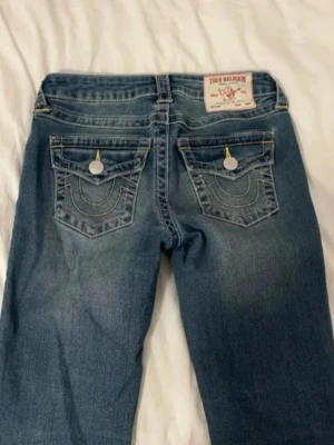 Blå jeans från True Religion - Säljer ett par blå jeans från True Religion med klassiska kontrastsömmar och snygga fickdetaljer bak. Jeansen har raka ben och coola knappar på fickorna. Materialet är denim i bomull och passformen är avslappnad men ändå stilren. Nya🫶🏼