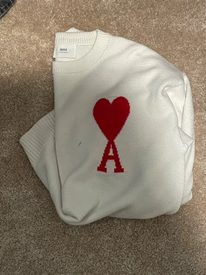 Vit stickad tröja från Ami Paris - Vit stickad tröja från Ami Paris med rött hjärta och bokstaven A på bröstet. Tröjan har rund halsringning, långa ärmar och ribbstickade muddar. Perfekt för dig som gillar stilrena och trendiga plagg.