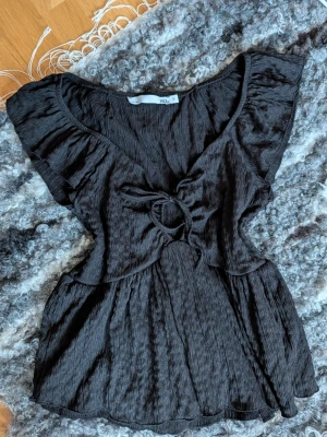 Svart topp med knytband från 157 - Supersnygg svart topp från 157 med knytband framtill och volang vid ärmarna. Knappt använd eftersom den har blivit lite för liten för mig :)