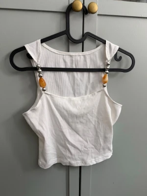 Vit ribbad linnetopp med pärldetaljer - Säljer en vit oandvänd ribbad linnetopp med breda axelband och snygga pärldetaljer i silver och bärnstensfärg framtill. Från SHEIN.  Toppen är croppad och har en enkel, clean look som passar perfekt till jeans eller kjol.