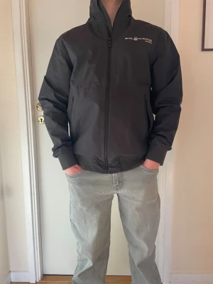 Svart Sail Racing vindjacka Gore-Tex - Snygg svart vindjacka från Sail Racing med hög krage och dragkedja framtill. Jackan har diskret logga på bröstet och Gore-Tex-detalj på ryggen. Tillverkad i slitstarkt material som står emot vind och regn. Perfekt för dig som vill ha en sportig och stilren look.  Fick den i julklapp men ville inte ha den använd max 5 gånger 