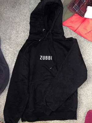Svart hoodie ZUBBI från Dunder Kläder - Säljer en svart hoodie från Dunder Kläder med texten 'ZUBBI' tryckt på bröstet. Hoodien har klassisk huva med dragsko och stor magficka framtill. Tillverkad i mjukt material, perfekt för chill dagar eller streetwear-stil.