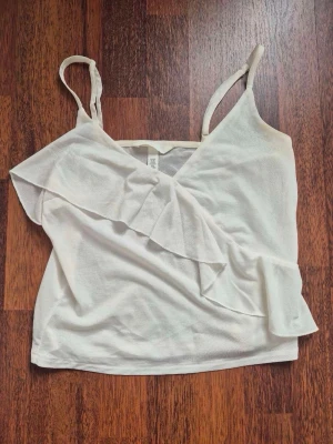 Vit volang topp från H&M - Säljer en söt vit linnetopp från H&M med tunna axelband och volangdetalj framtill. Toppen har en v-ringad skärning och är perfekt för varma dagar. Enkel att matcha med jeans eller kjol för en fräsch look.