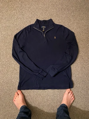 Mörkblå half-zip tröja Polo Ralph Lauren - Mörkblå långärmad tröja från Polo Ralph Lauren med klassisk half-zip och liten broderad logga på bröstet. Tröjan har hög krage och är tillverkad i mjukt bomullsmaterial. Perfekt för en avslappnad men stilren look.