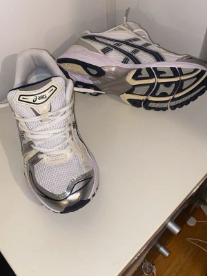 Vita ASICS sneakers med silverdetaljer - Snygga vita sneakers från ASICS med mesh-ovandel och silvriga detaljer. Skorna har svarta och grå inslag samt en chunky sula för extra komfort. Klassisk snörning och sportig design som passar perfekt till streetwear och casual outfits.