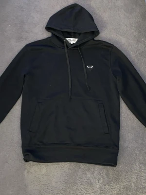 Svart hoodie från Comme des Garçons Play - Säljer en svart hoodie från Comme des Garçons Play med den ikoniska hjärtloggan med ögon på bröstet. Hoodien har huva med dragsko och en stor magficka. Tillverkad i mjuk bomull, perfekt för en avslappnad streetwear-look.