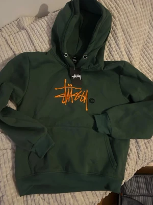 Grön Stüssy hoodie med orange logga - Snygg grön hoodie från Stüssy med stor orange broderad logga på bröstet och svart copyright-märke. Klassisk känguruficka framtill och justerbar huva med snören. Perfekt för dig som gillar streetwear och vill sticka ut lite extra.