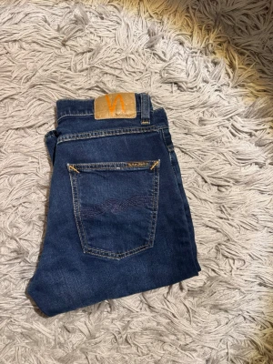 Nudie Jeans - Säljer ett par mörkblå jeans från Nudie Jeans med klassisk femficksmodell och gul kontrastsöm. Jeansen har rak passform och är tillverkade i slitstark denim med Nudie-logga på bakfickan. Perfekta för dig som gillar stilrena och tidlösa jeans. De är i 10/10 skick och bara att höra av sig vid minsta fundering! 