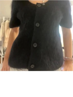 Svart fluffig kofta från COS - Kortärmad svart kofta från COS i fluffigt material med stora knappar framtill och v-ringad hals. Perfekt för lager på lager och ger en trendig look. Materialet ser ut att vara en mjuk blandning med mohair eller liknande. Använd men i bra skick storlek M. Hör gärna av er vid frågor pris går alltid att diskuteras💕