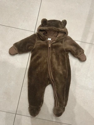 Brun teddyoverall med öron - Supersöt brun teddyoverall med huva och små björnöron. Dragkedja framtill och mjukt, fluffigt material som håller värmen. Overallen har vikbara muddar vid ärmar och ben för extra mysfaktor. Perfekt för kyliga dagar! Jätte söt overall   Dragkedja med halsskydd   Pile   Trikåfodrad   Stygnen har sprätts upp lite vid halsen, går lätt att sy. Se bild