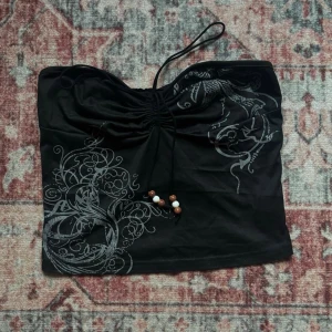 Svart tubtopp med snörning och pärlor - Snygg svart tubtopp utan axelband, snörning framtill och dekorativa pärlor. Toppen har ett grått mönster med detaljerade tryck på framsidan. Är ganska kort.