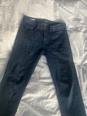 Mörkblå jeans från Jack & Jones  - Säljer ett par mörkblå jeans från Jack & Jones med raka ben och diskreta slitningar framtill. Klassisk femficksmodell med dragkedjagylf och med Jack & Jones logga bak i midjan. Tillverkade i bomull med en snygg tvättad finish.