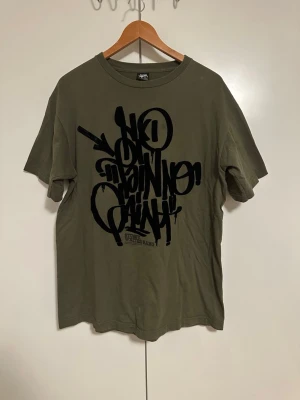 Olivgrön Stüssy t-shirt med tryck - Säljer en olivgrön t-shirt från Stüssy med stort svart graffiti-tryck på framsidan. Klassisk rund hals och korta ärmar. Tillverkad i mjuk bomull som känns skön mot huden. Perfekt för dig som gillar streetwear och vill sticka ut.