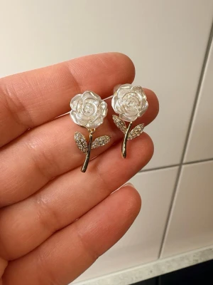 Blommiga örhängen med pärldetaljer - Snygga örhängen i form av vita rosor med pärlliknande material och glittrande blad. Stjälken är i guldfärg och bladen är dekorerade med små stenar för extra bling. Perfekt för dig som vill ha något unikt och sött till din stil.