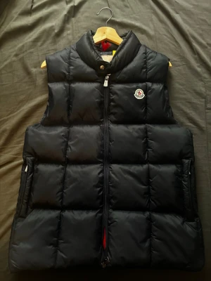 Mörkblå dunväst från Moncler - Snygg mörkblå dunväst från Moncler med hög krage och dragkedja framtill. Västen har Moncler-logga på bröstet och två fickor med dragkedja samt en innerficka. Baksidan har coola detaljer i vitt och rött längst ner. Perfekt för lager-på-lager och streetstyle. Pris går att diskutera, skriv gärna om du har några frågor!💙