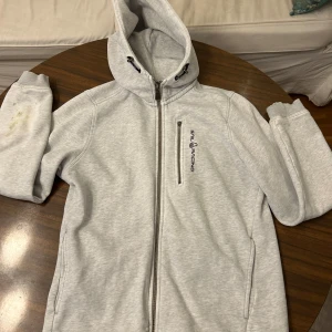 Ljusgrå hoodie från Sail Racing - Ljusgrå hoodie från Sail Racing med hel dragkedja och huva med snörning. Tröjan har en bröstficka med dragkedja och Sail Racing-logga på framsidan. Mjuk insida i fleece och långa ärmar med ribbade muddar. Perfekt för chill eller utomhus. Den har en rätt så stor fläck syns inte så mycket! På armen