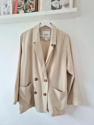Beige kavaj från Monki - Stilren beige kavaj från Monki med dubbelknäppning och stora bruna knappar. Kavajen har två rymliga framfickor och en avslappnad passform. Perfekt för en trendig och enkel look. Materialet ser mjukt och bekvämt ut.