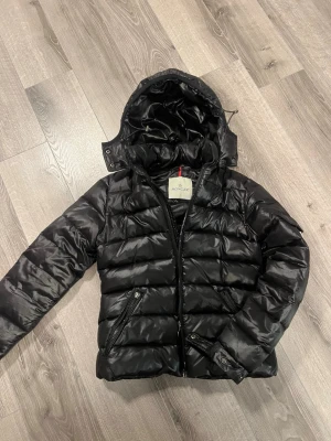 Monclear jacka herr  - Snygg svart dunjacka från Moncler med avtagbar huva och dragkedja framtill. Jackan har två snedställda fickor med dragkedja och quiltad design. Perfekt för kalla vinterdagar och riktigt stilren look. Ena jackan har en söndrig dragkedja annars bra skick 