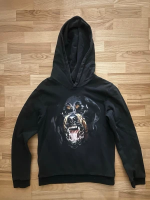 Svart Givenchy hoodie med hundmotiv - Säljer en svart Givenchy hoodie med stor huva och ett fett tryck av en morrande rottweiler på bröstet. Hoodien har ribbade muddar vid ärmslut och nederkant. Perfekt för dig som vill sticka ut med en cool och edgy stil.
