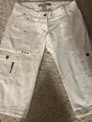 Vita cargo shorts från Blue Seven - Säljer ett par vita cargo shorts från Blue Seven med flera fickor, dragkedjedetalj och knappstängning. Shortsen har en avslappnad passform och är perfekta för sommaren. Tillverkade i bomull för en skön känsla.
