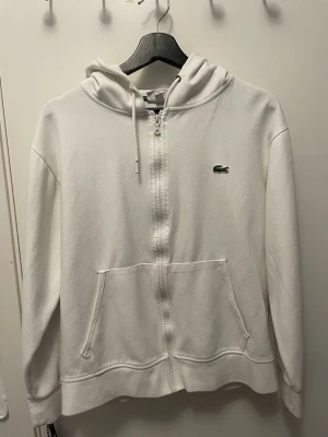 Vit zip hoodie från Lacoste - Klassisk vit hoodie från Lacoste med dragkedja framtill och broderad grön krokodil på bröstet. Hoodien har huva med dragsko och en stor magficka. Tillverkad i mjukt bomullsmaterial och har ribbade muddar vid ärmslut och nederkant.