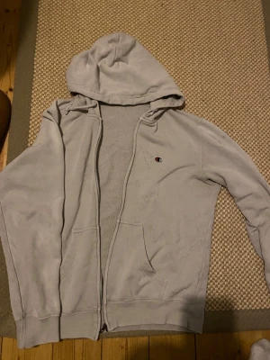 Ljusgrå zip hoodie från Champion - Ljusgrå hoodie från Champion med dragkedja framtill och två fickor. Klassisk huva och liten Champion-logga broderad på bröstet. Mjuk insida i fleece och avslappnad passform. Perfekt för chill dagar eller till träningen.