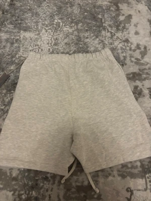 Grå shorts från Essentials Fear of God - Snygga ljusgrå shorts från Essentials Fear of God med elastisk midja och snörning. Tillverkade i mjuk bomull för maximal komfort. Minimalistisk design med logga tryckt på benet och avslappnad passform.