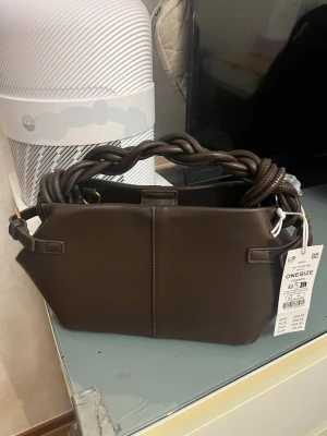 Brun handväska med flätat handtag - Snygg brun handväska från GINATRICOT. Den är helt ny, men se bild 2 och 3. Den var så när jag köpte. Helt slutsåld på hemsidan. 