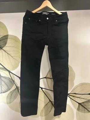 Svarta Dressman Jeans regular fit - Snygga svarta regular fit jeans från Dressman med klassisk femficksdesign och knappgylf. Jeansen har raka ben och är tillverkade i ett stretchigt bomullsmaterial för skön passform. Perfekta till alla dina outfits!