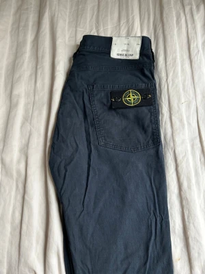 Mörkblå chinos från Stone Island - Säljer ett par mörkblå byxor från Stone Island med klassisk patch på bakfickan. Byxorna har raka ben och normal passform, tillverkade i mjuk bomull. Perfekta för dig som gillar streetwear och vill ha något stilrent men ändå med edge.