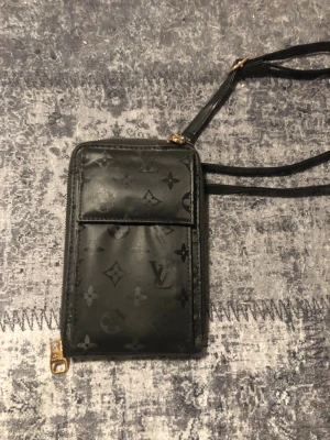 Svart Louis Vuitton axelväska - Snygg svart handväska från Louis Vuitton med det klassiska monogrammönstret. Väskan har guldfärgade detaljer, smal axelrem och dragkedja. Tillverkad i läderliknande material och har en rektangulär form som ger en modern och exklusiv känsla.