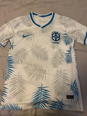 Vit Brasilien fotbollströja Nike utan namn o nummer - Snygg vit Brasilien fotbollströja från Nike med blå och grå palmbladsmönster. Tröjan har blå detaljer vid krage och ärmslut, samt Brasilien-logga och Nike swoosh på bröstet. Perfekt för dig som vill sticka ut på planen eller heja på landslaget.
